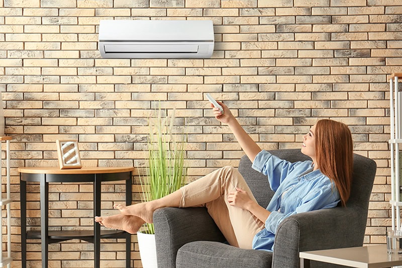 How to Cool the Upstairs of a Two-Story Home. Young woman switching on air conditioner while sitting in armchair at home.